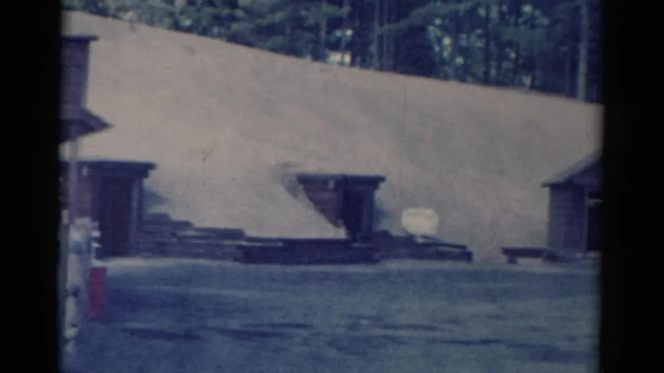 1959 Catskill-area 8mm frame showing a pale sloped outdoor embankment, small wooden structures, dark rails, and a wooded background.