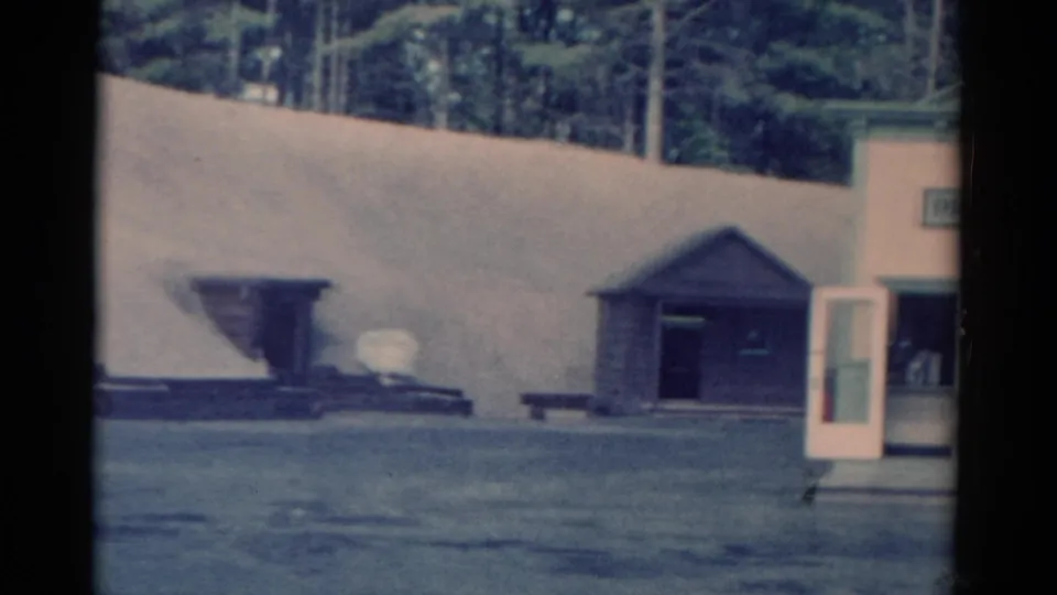 1959 Catskill-area home-movie frame showing a small wooden shed, open booth door, pale embankment, and sparse outdoor grounds.