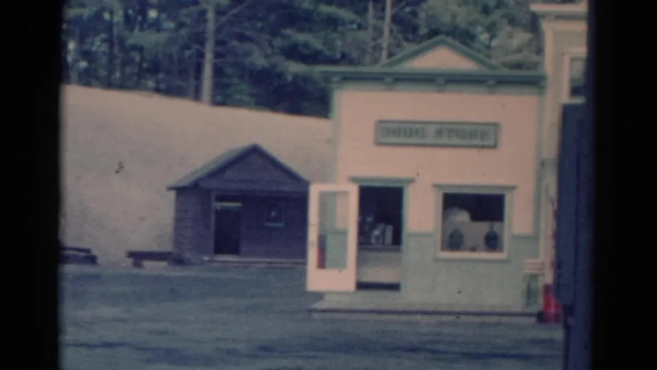 1959 Catskill-area 8mm frame showing a light-colored drug store exterior with signage, open doorway, display window, and adjacent shed.