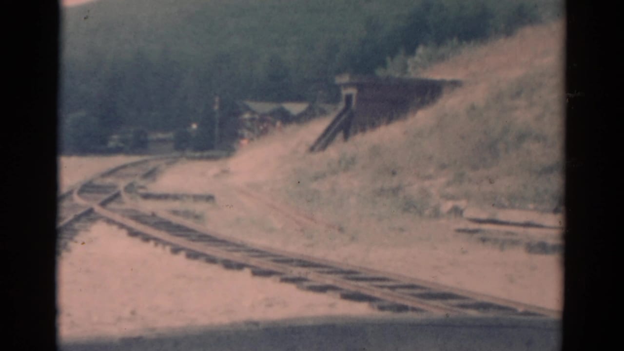 1959 Catskill-area 8mm frame showing a red train approaching on curving tracks near a small trackside structure.