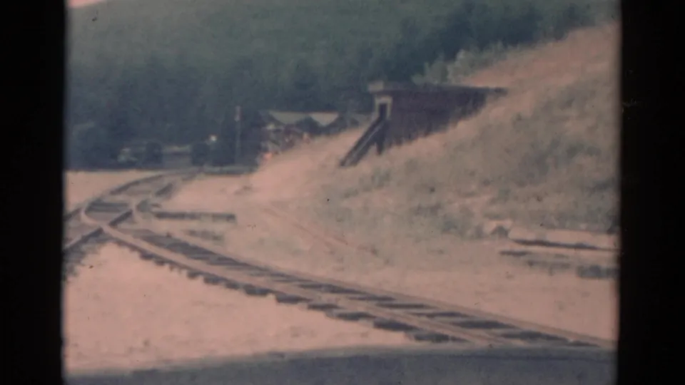 1959 Catskill-area 8mm frame showing a red train approaching on curving tracks near a small trackside structure.