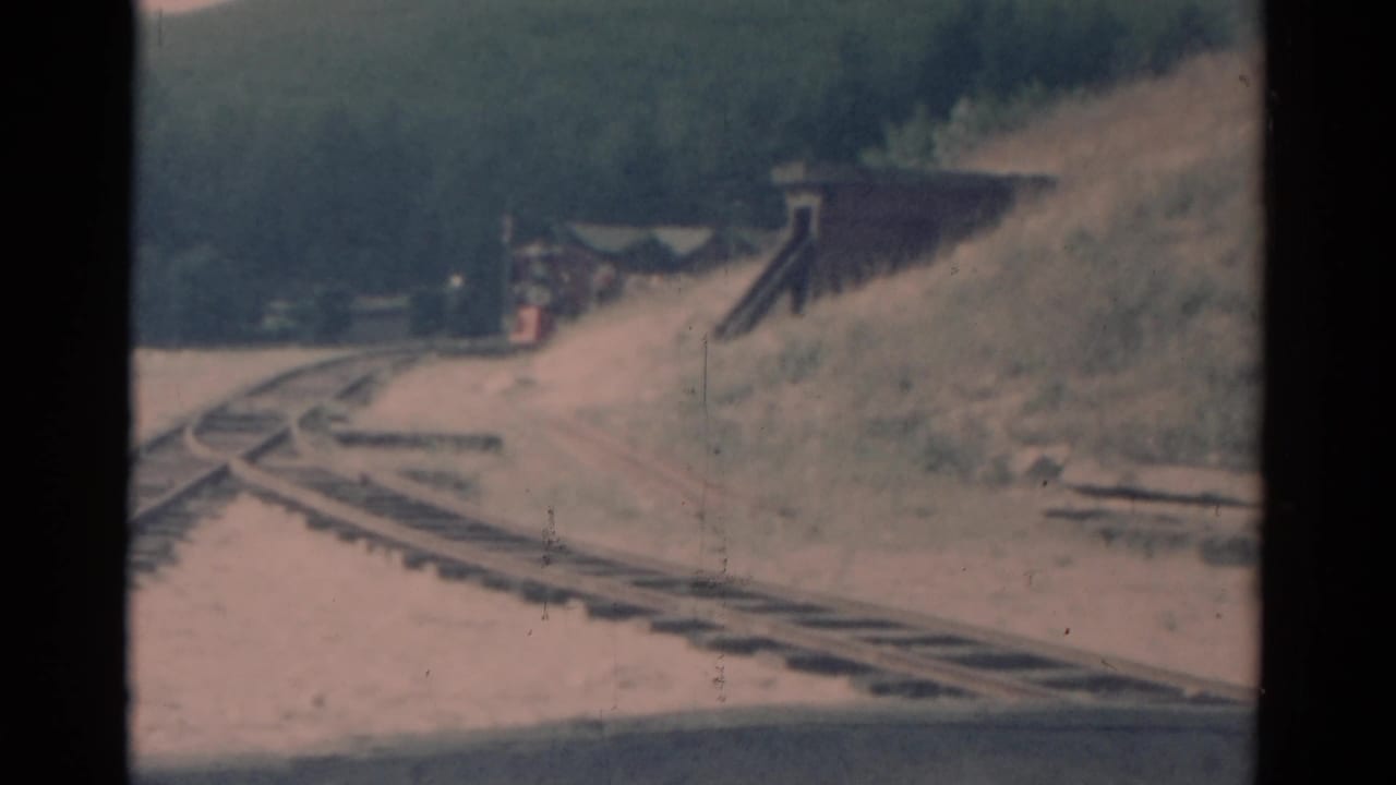 1959 Catskill-area home-movie frame with a train moving closer beside a grassy embankment and trackside structure.