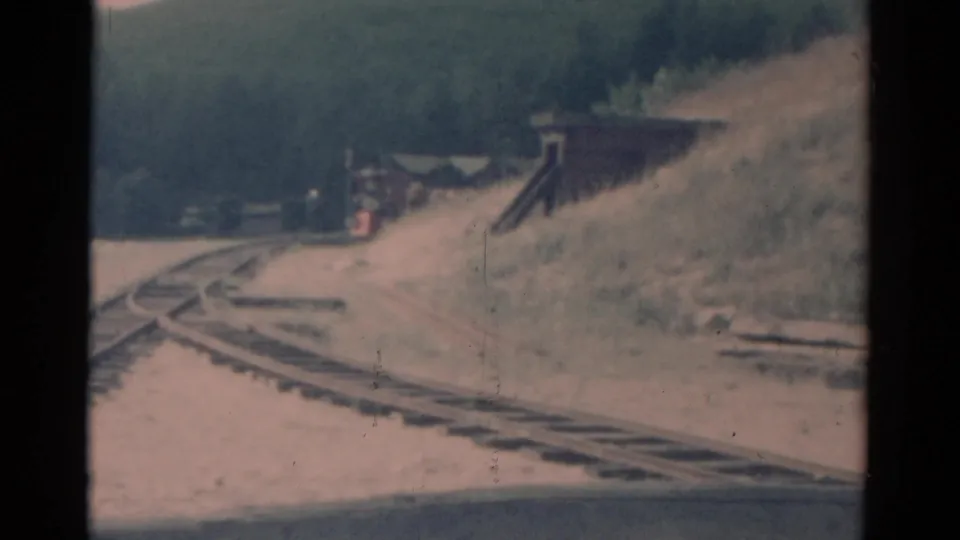 1959 Catskill-area home-movie frame with a train moving closer beside a grassy embankment and trackside structure.