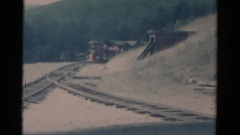 1959 Catskill-area frame of a red train rounding the mountain curve near a small trackside building.