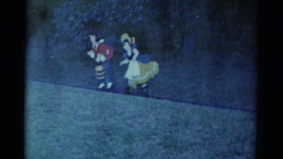 1959 Catskill frame of two costumed walkaround characters, one resembling Snow White, moving across grass near trees at night.