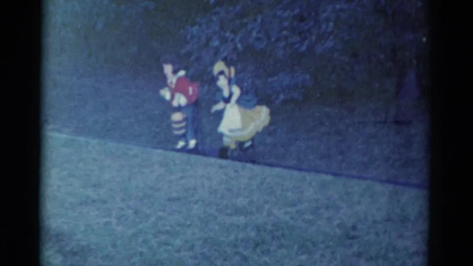 In Catskill, New York, 1959 home-movie footage, two costumed performers move along a grassy path near dark foliage.