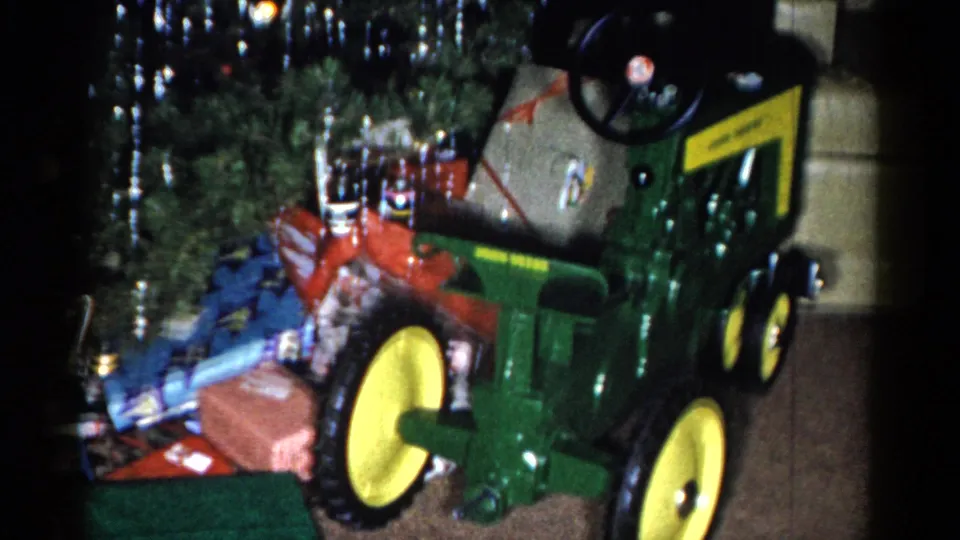 A closer 1959 8mm frame shows the toy tractor dominating the foreground with wrapped Christmas gifts and tree decorations behind it.