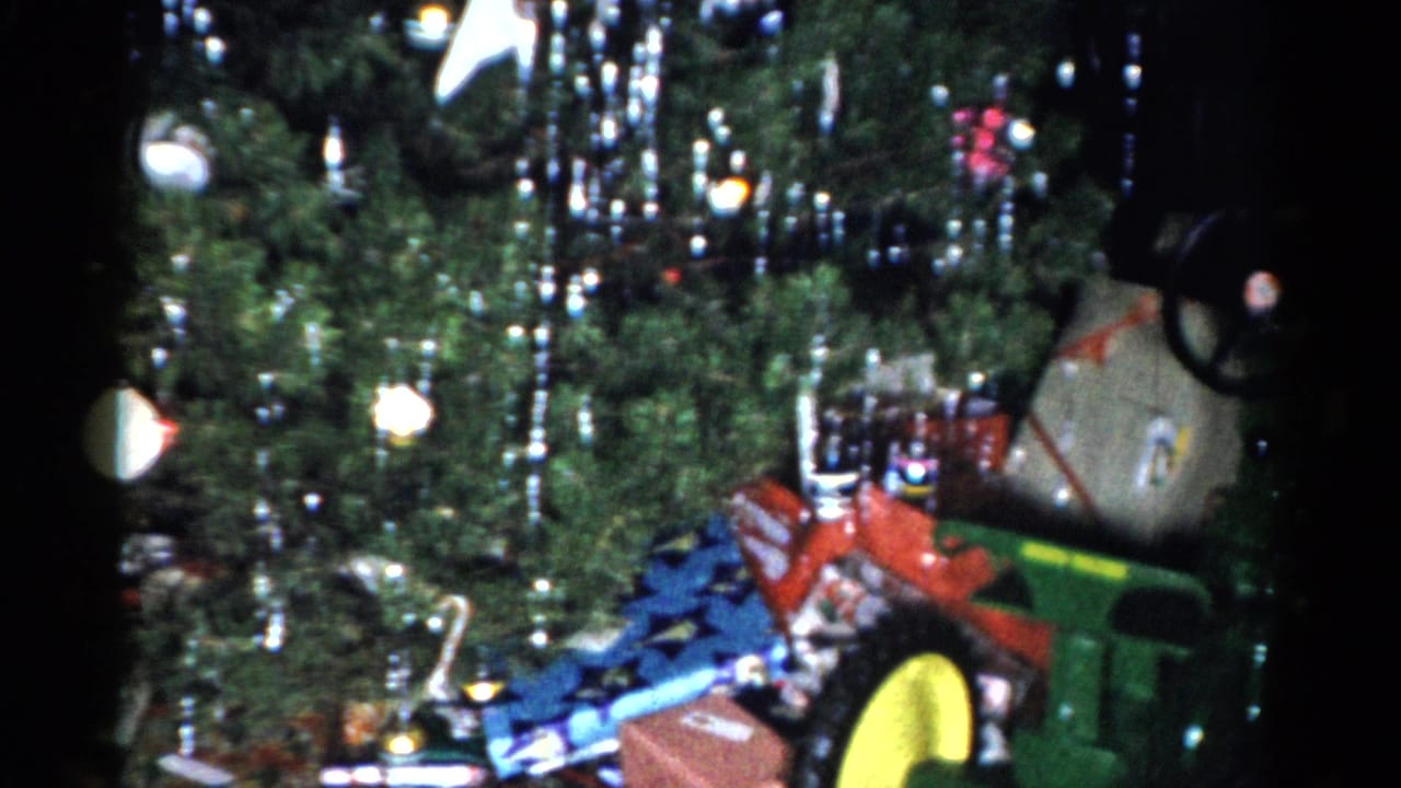 1959 Catskill home-movie frame of a Christmas tree with silver tinsel, white ornaments, presents, and a green toy tractor among the gifts.