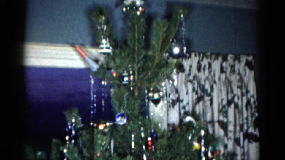 1959 Catskill home-movie frame of the Christmas tree near patterned curtains, showing sparse branches, tinsel, ornaments, and interior wall trim.