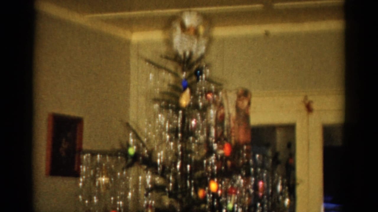 1959 Catskill home-movie frame showing a Christmas tree with tinsel, lights, ornaments, a wall picture, and a cream doorway.