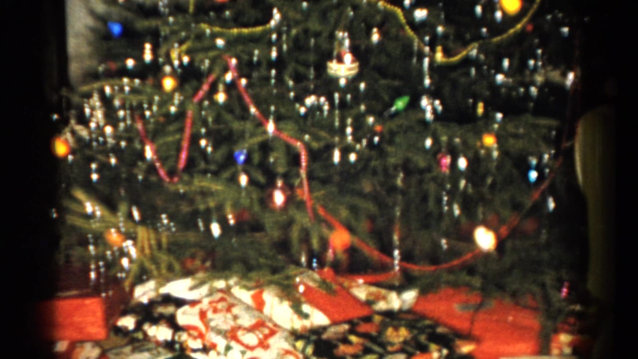 1959 Catskill home-movie frame showing a Christmas tree with colored bulbs, tinsel, red garland, and gifts on the floor.