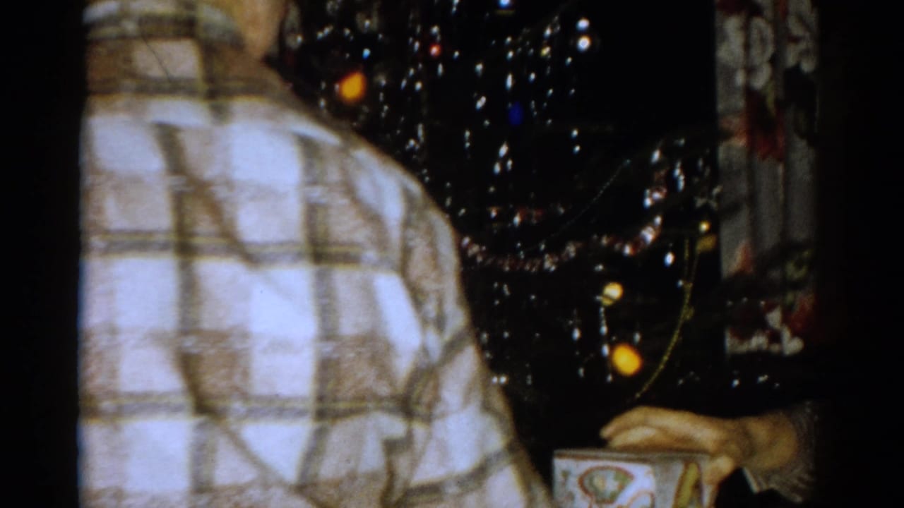 In a 1959 Catskill, New York home movie, a person in a plaid shirt stands near a decorated Christmas tree with tinsel and ornaments.