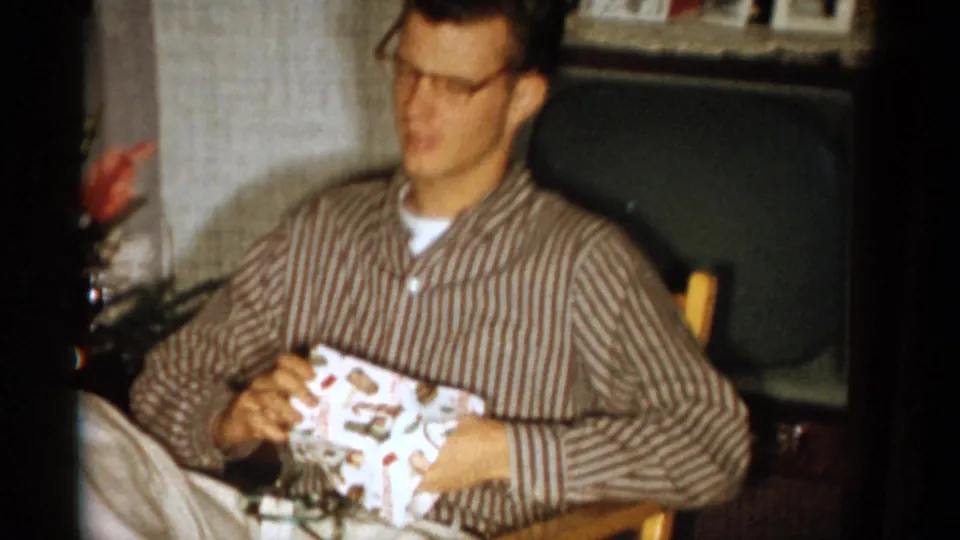 A young man in glasses and a striped shirt opens a wrapped gift in a 1959 Catskill living room, with a television behind him.