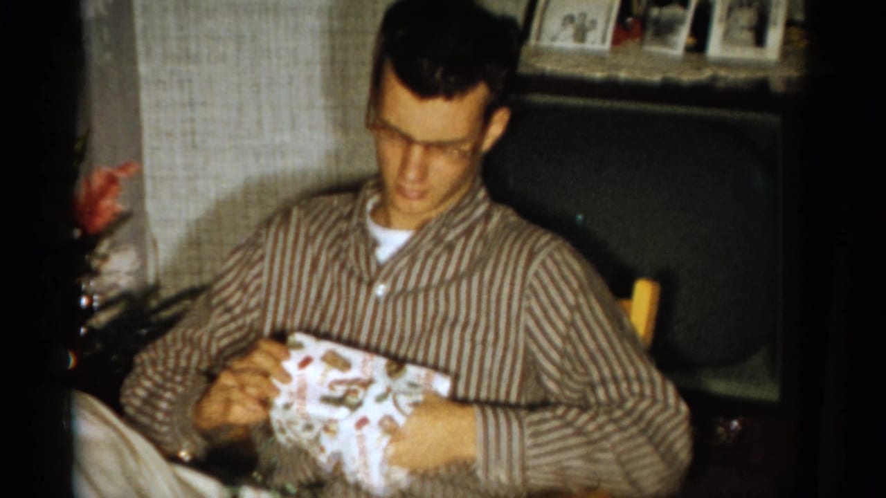 Bespectacled man in striped pajamas unwraps a Christmas gift indoors.
