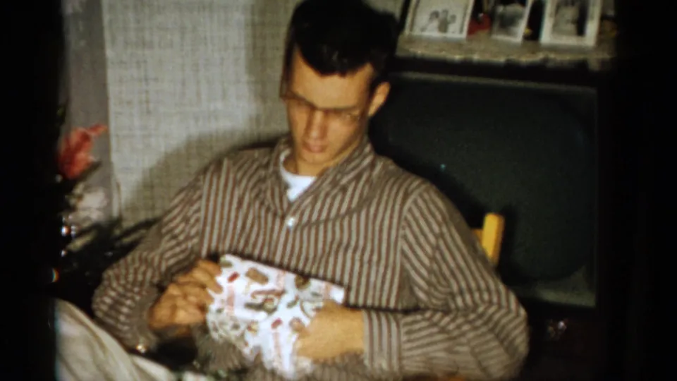 In a Catskill, New York home-movie frame from 1959, a bespectacled young man in striped pajamas opens patterned wrapping paper beside a chair.