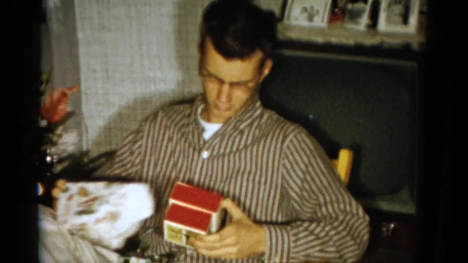 The 1959 Catskill home-movie frame shows the man lifting a red-and-white gift box while holding loosened wrapping paper.