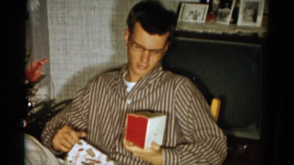 A bespectacled man in striped pajamas holds the opened gift box in a private Catskill interior, with framed photos and furniture behind him.