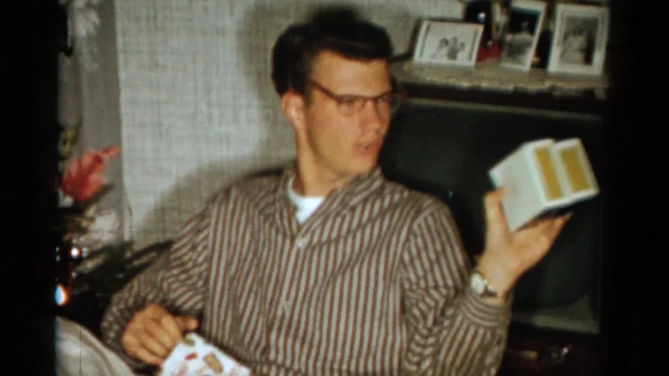 The man turns toward someone off camera and raises the opened box, with a vintage television and framed photographs visible in the home interior.