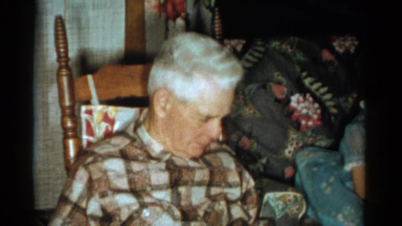 Elderly white-haired man in a checkered shirt seated in a wooden rocking chair.