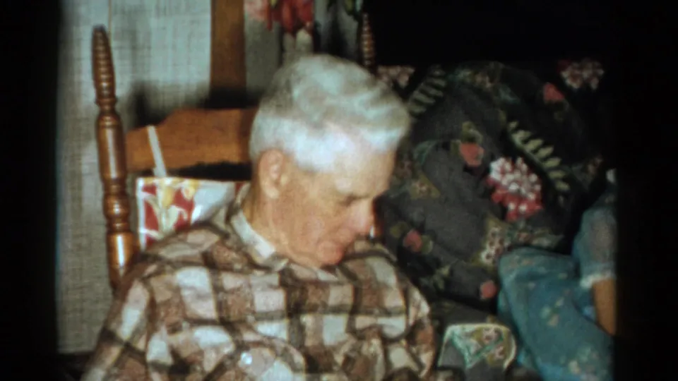 In 1959 Catskill home-movie footage, an elderly white-haired man sits in a wooden rocking chair beside floral-patterned upholstery.