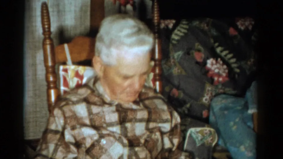 The elderly man in the checkered shirt looks downward in a Catskill, New York private home interior filmed in 1959.