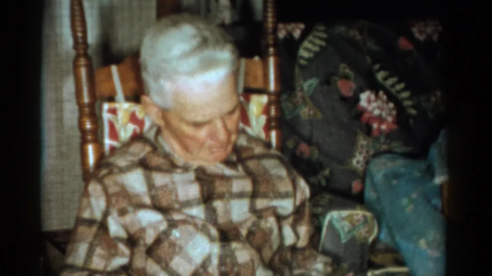 An elderly man with white hair sits with his head lowered, framed by patterned upholstery and curtains in 1959 home-movie footage.