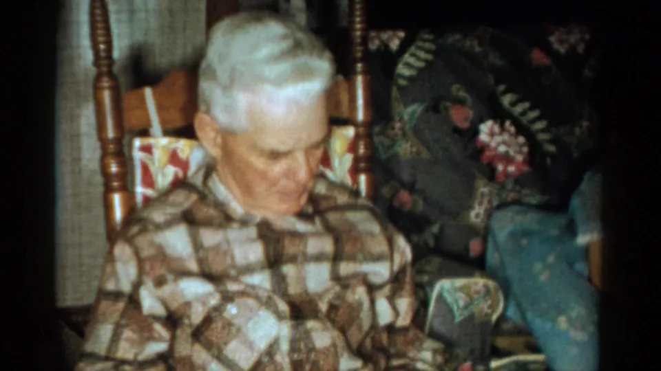 The man remains seated in the wooden rocking chair, with floral upholstery and curtains visible in the 1959 Catskill interior scene.