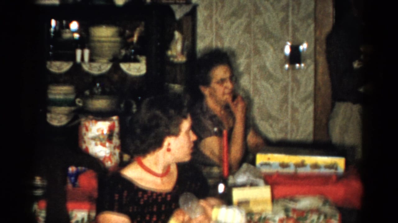 Two women sit at a holiday table with wrapped gifts and a china cabinet behind them.