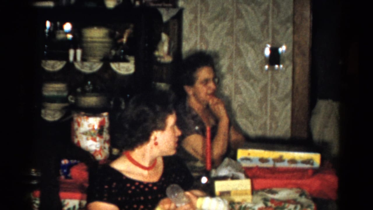 Two women remain seated at the table, with gifts, dishes, and patterned wallpaper visible.