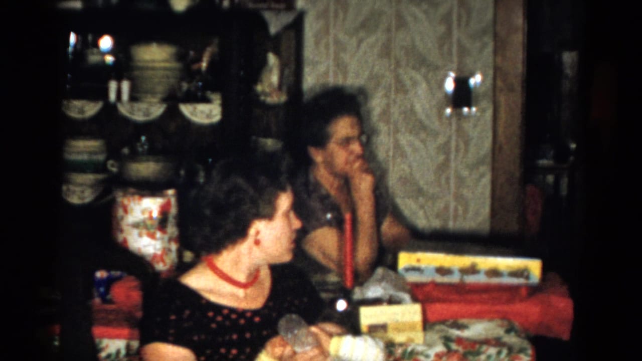 Home-movie frame from the Catskill Christmas gathering, showing a woman in a dark dress turned at the table as another woman sits near the wall.