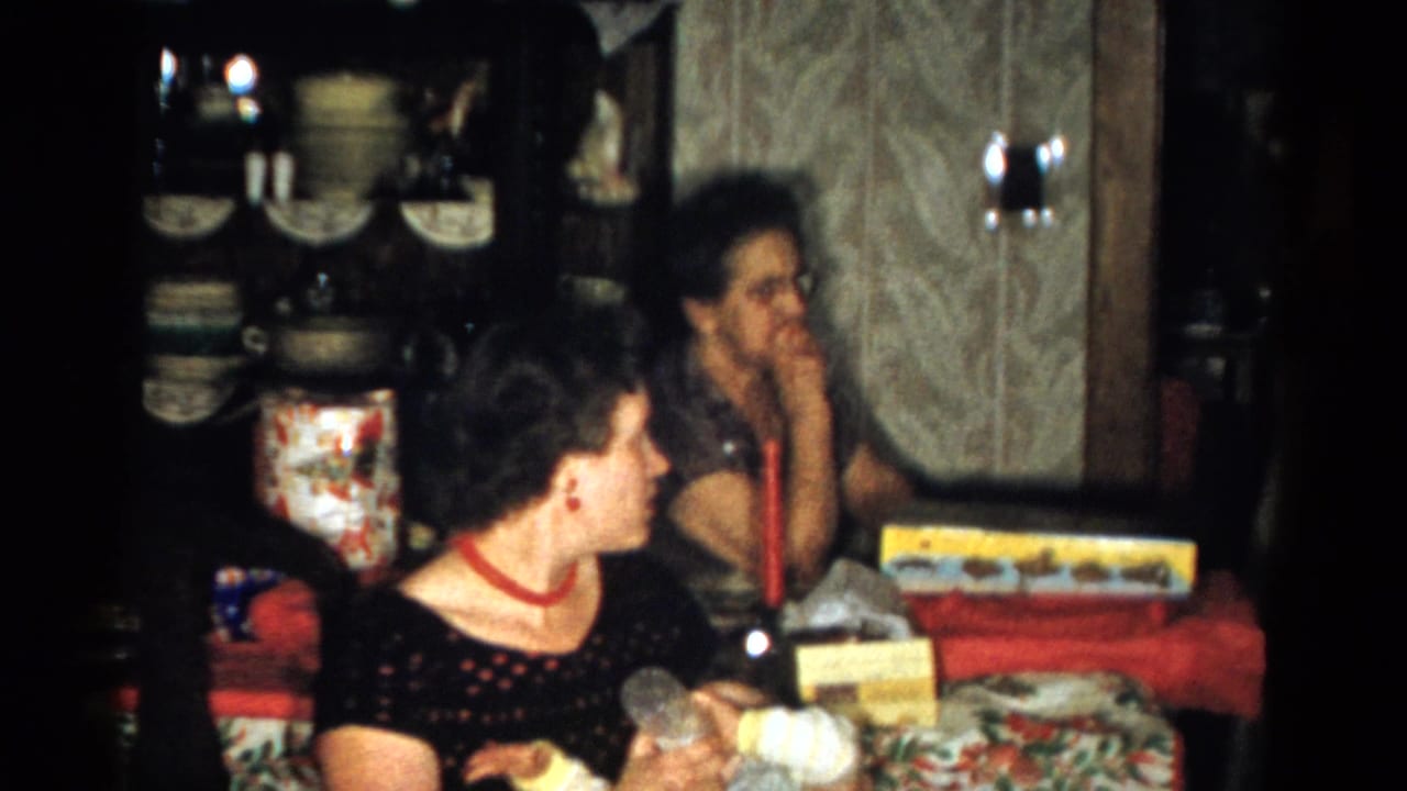 1959 Catskill Christmas interior showing two women at a red-covered table with presents, a candle, patterned wallpaper, and a dish cabinet.
