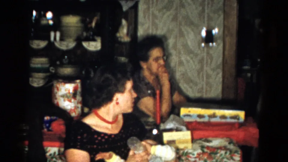 1959 Catskill Christmas interior showing two women at a red-covered table with presents, a candle, patterned wallpaper, and a dish cabinet.