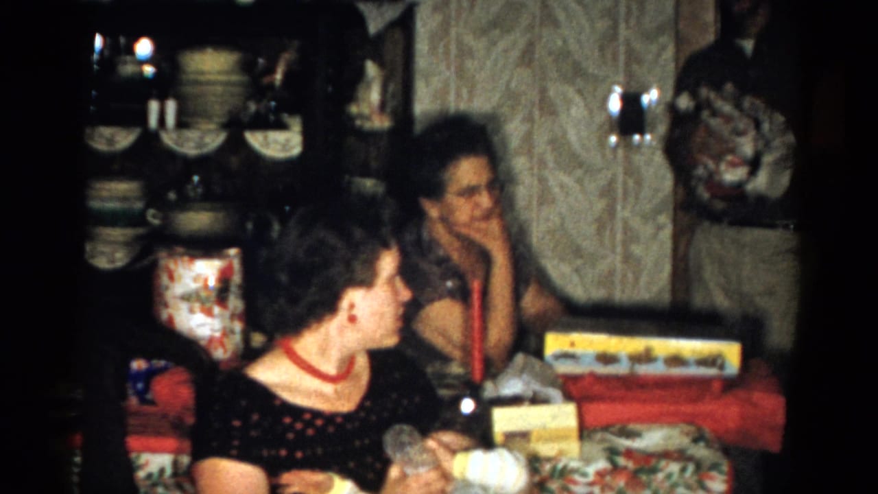 Adults sit and stand around a table with wrapped gifts in a dim living room.