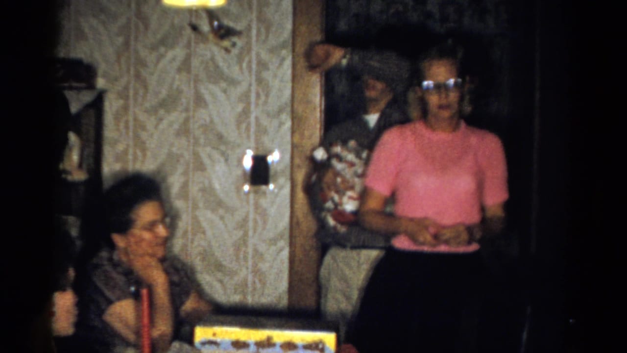 Catskill, New York home-movie frame from 1959 showing a standing woman in pink near a seated woman, gifts, and patterned wallpaper.
