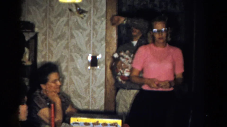 Catskill, New York home-movie frame from 1959 showing a standing woman in pink near a seated woman, gifts, and patterned wallpaper.