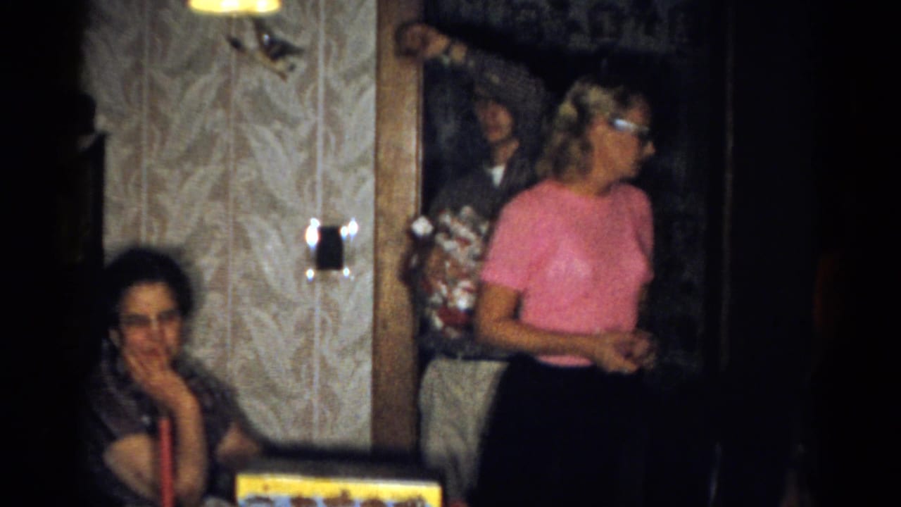 1959 Catskill home-movie frame with a woman in pink turned toward the room, a seated woman, gift box, red candle, and doorway figures.