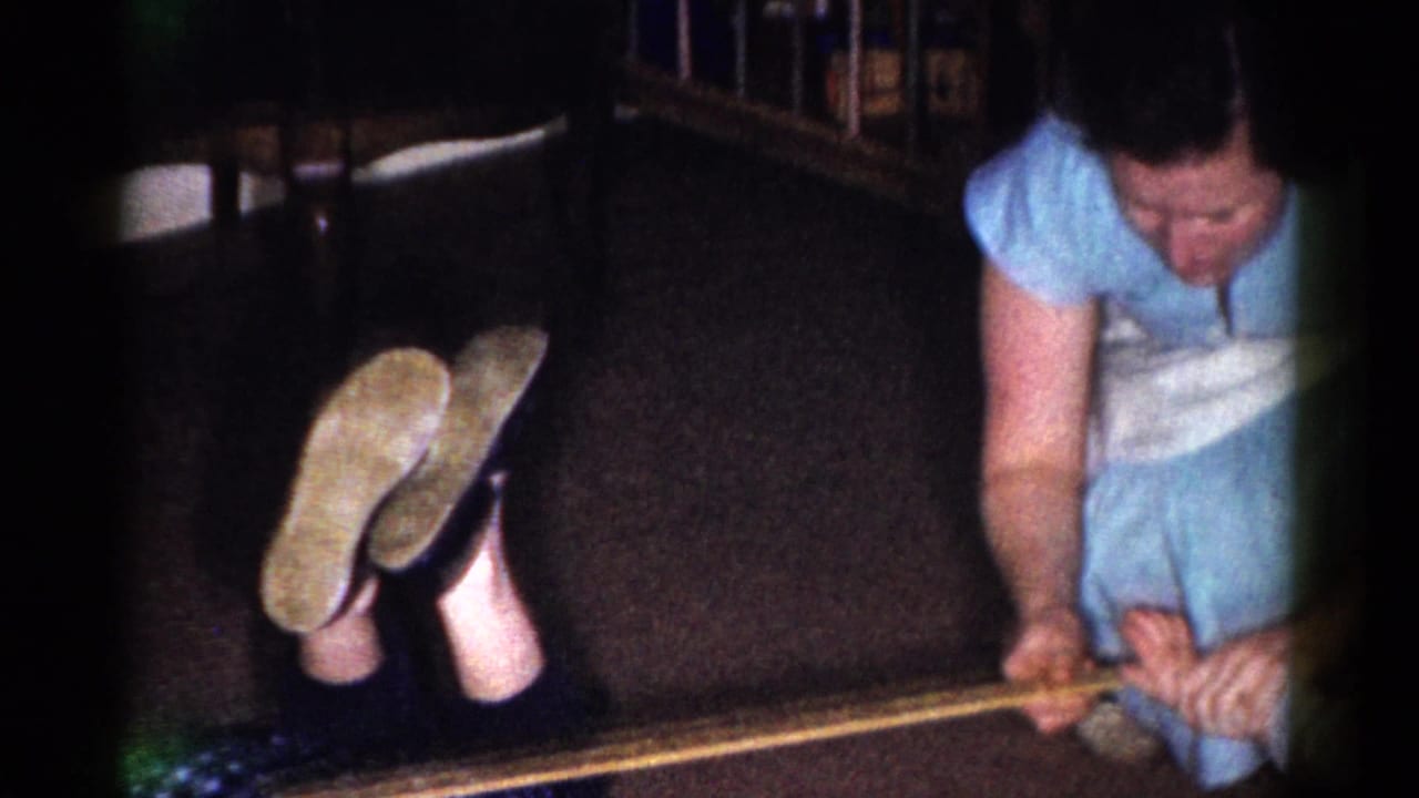 Woman in a blue dress and white apron crouches near a wooden yardstick on a dark floor.
