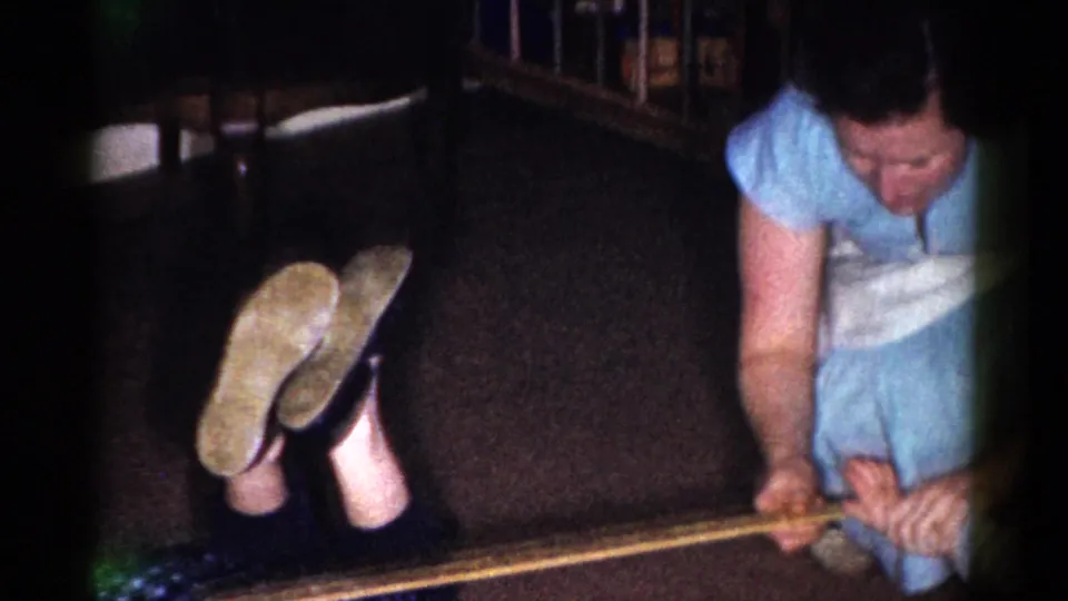 In a 1959 Catskill home-movie frame, a woman in a blue dress and white apron crouches beside a yardstick while another person's legs appear nearby.