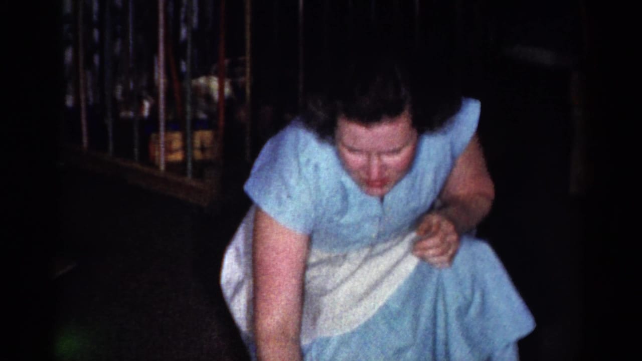 In the 1959 home-movie frame, the woman crouches in her blue dress and white apron, reaching forward near the floor in a private interior.