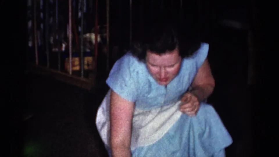 In the 1959 home-movie frame, the woman crouches in her blue dress and white apron, reaching forward near the floor in a private interior.