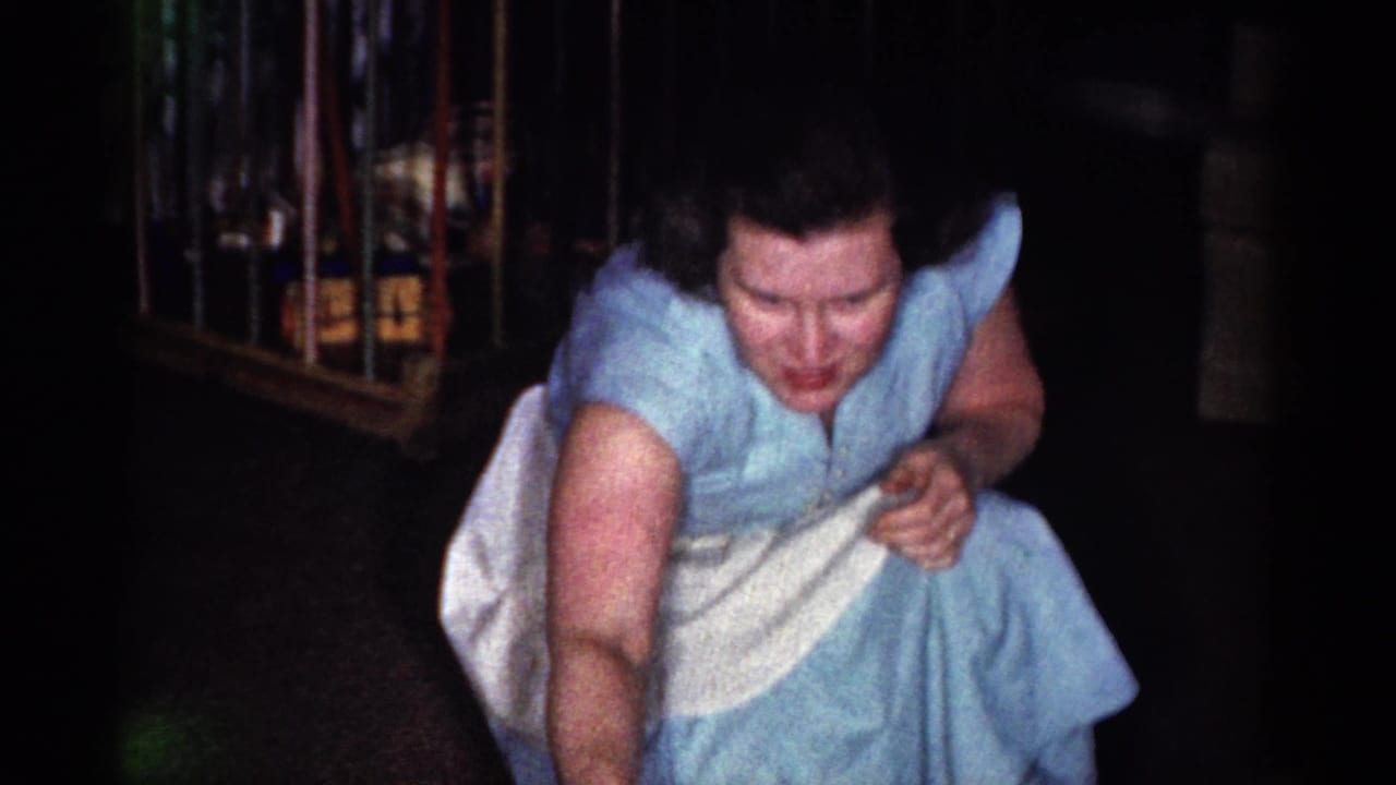 The woman leans forward from a crouch in the Catskill home-movie interior, her face visible above a white apron and blue dress.