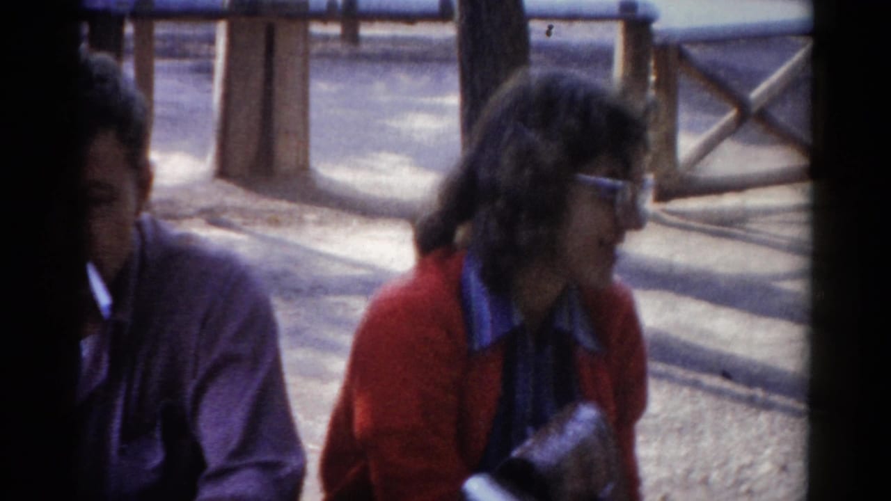 Two adults sit outdoors near wooden railings, with the woman in a red cardigan and the man partly visible.