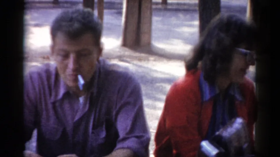 A 1959 Catskill home-movie frame shows the man smoking with his head lowered as the woman in red sits nearby outdoors.