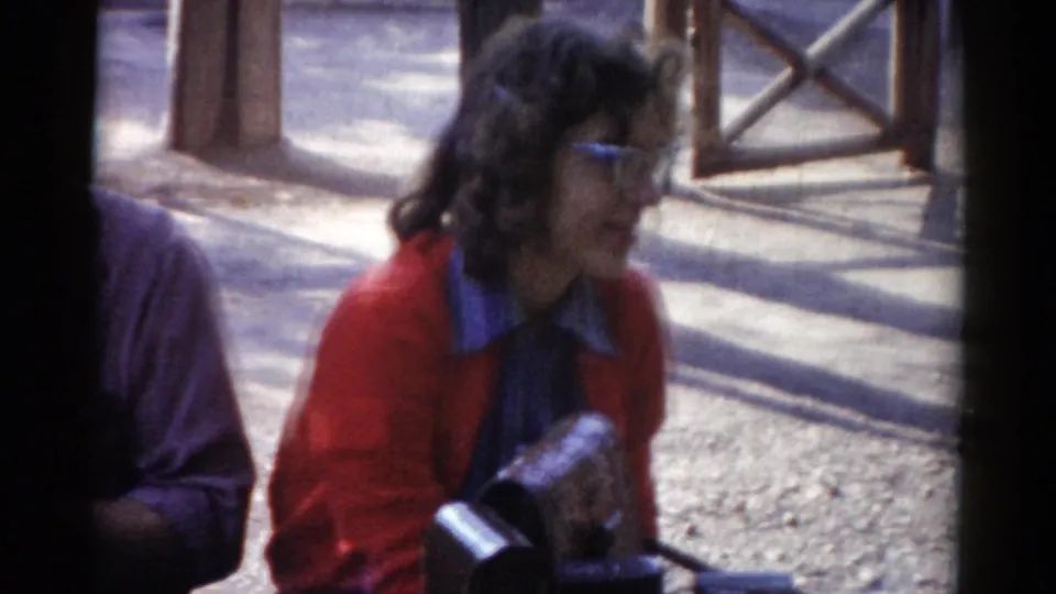 In this 1959 Catskill frame, the woman in a red cardigan and glasses sits in profile near wooden railings as the man is partly cropped.
