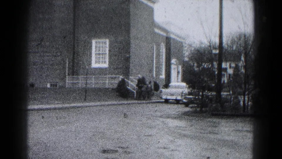 1959 Hagerstown home-movie frame with a brick building, parked car, bare trees, and distant people along a quiet street.