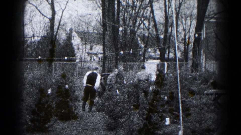 1959 Hagerstown backyard view with adults near small evergreens, bare trees, string lights, fencing, and neighboring houses in soft 8mm black-and-white.