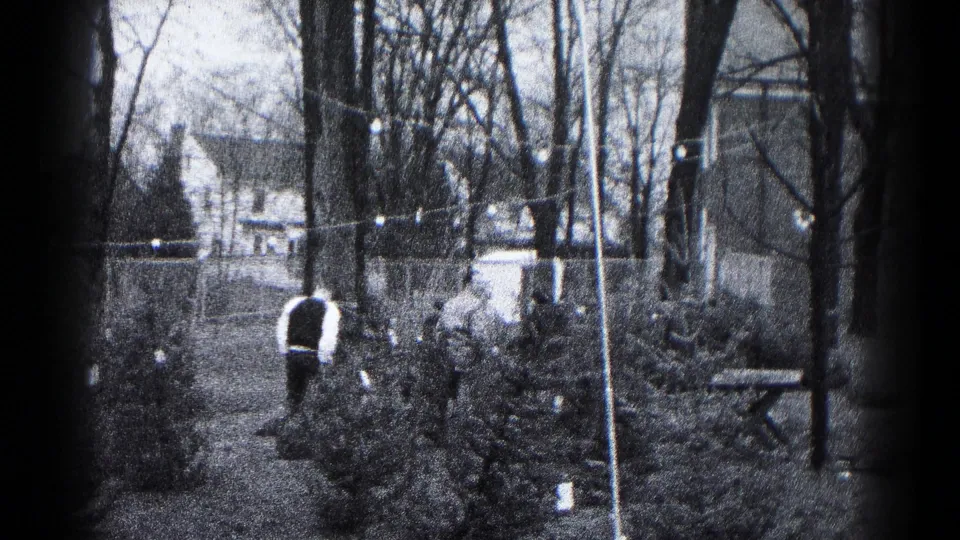 An adult walks away in a Hagerstown backyard, with small trees, bare branches, string lights, fencing, and a picnic table visible in the 1959 home movie.