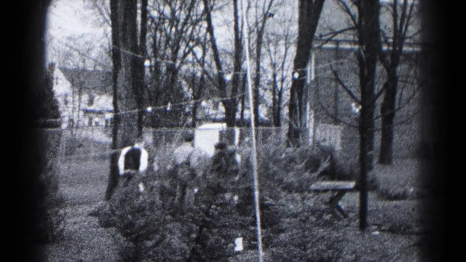 Adults appear to tend or inspect shrubs in a 1959 Hagerstown backyard, framed by bare trees, string lights, fencing, and distant houses.