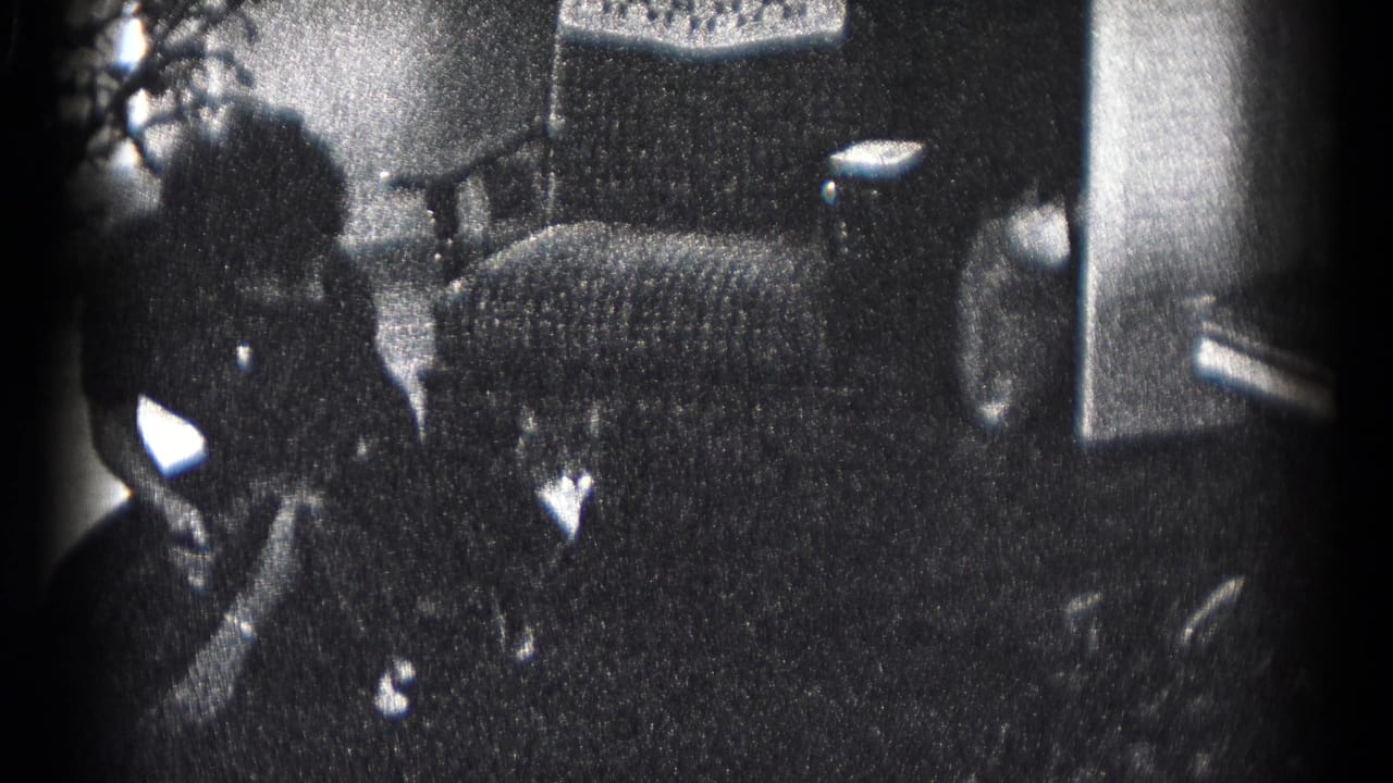 In 1959 Hagerstown home-movie footage, two adults stand indoors near an upholstered chair, plants, and small holiday decorations.