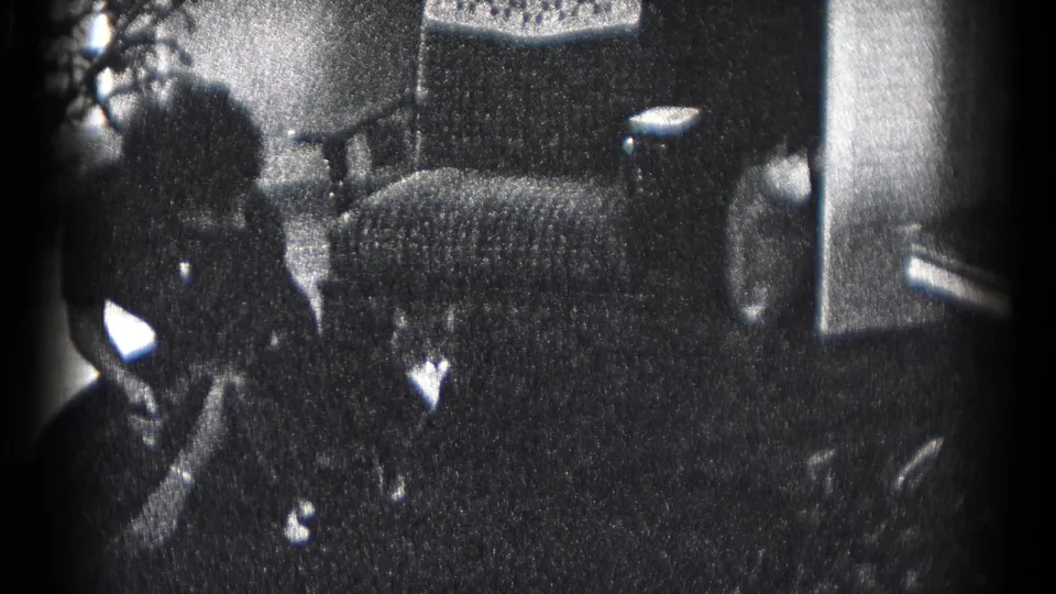In 1959 Hagerstown home-movie footage, two adults stand indoors near an upholstered chair, plants, and small holiday decorations.